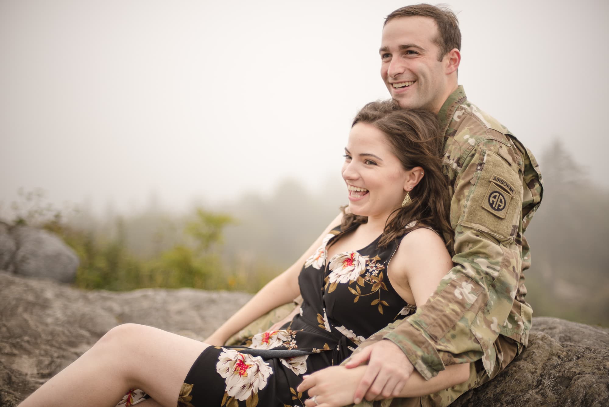 Military man sitting on mountain top with arms around fiance sitting on a foggy day near Asheville North Carolina photography done by Kathy Beaver.