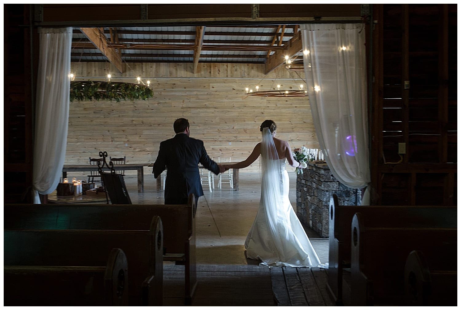 Bride and groom holding hands walking away from camera and into reception room inside beautiful wooden barn