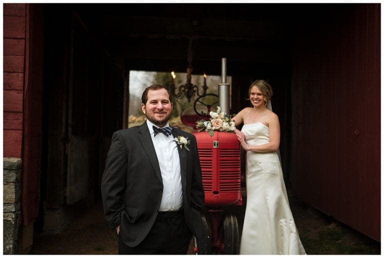 Bride and groom posing beside red rustic tractor, bride leaning against tractor smiling at groom while groom stands in front of tractor with hands in his pockets smiling and looking at camera