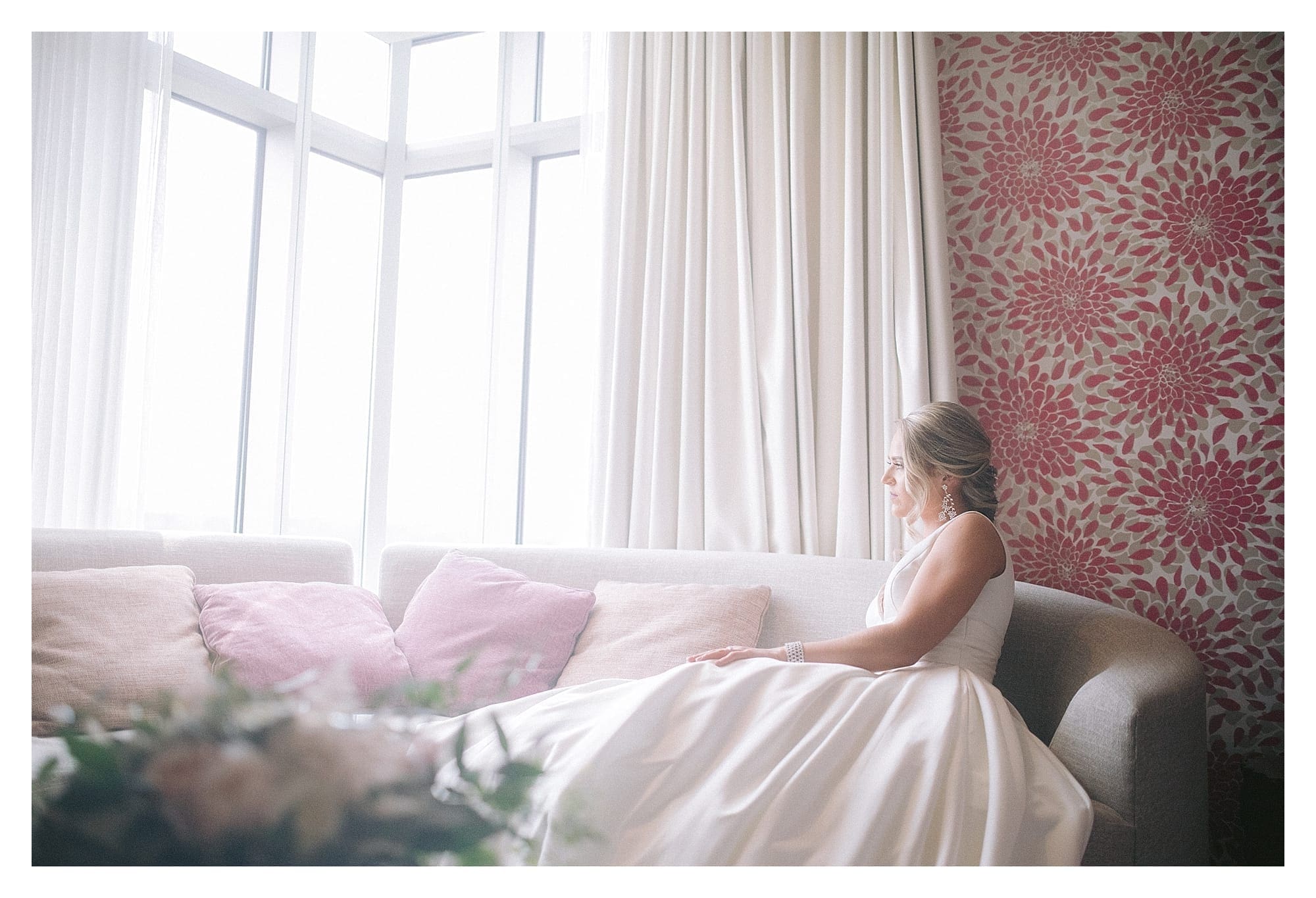 Bride in wedding dress sitting on couch looking out large window