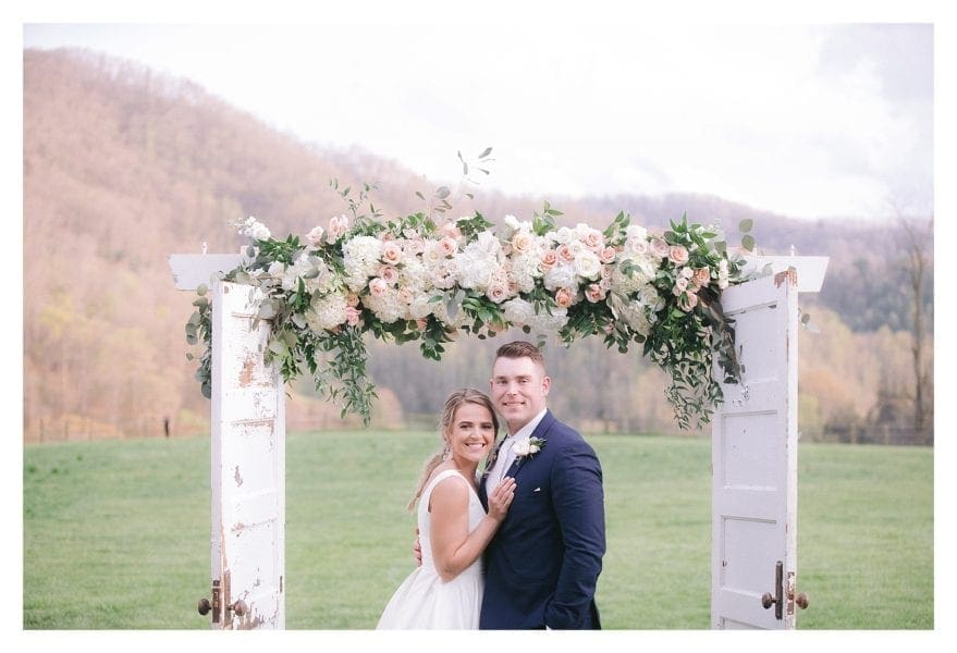 Bride and groom posing under wedding arbour of cream and peach flowers outdoors in grassy field