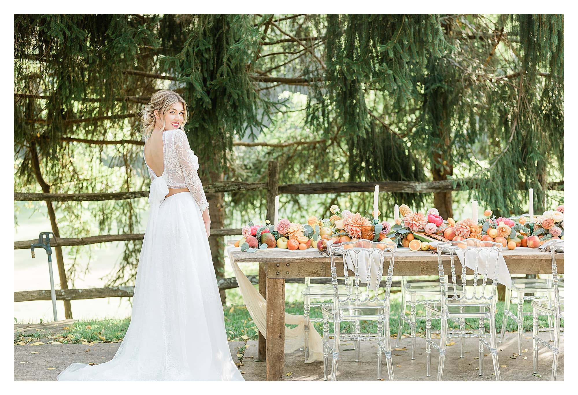 Rustic wooden table and clear plastic chairs with peach, pink and yellow citrus fruits as centrepiece down center of table with white candles with bride in white two piece lace gown standing beside smiling