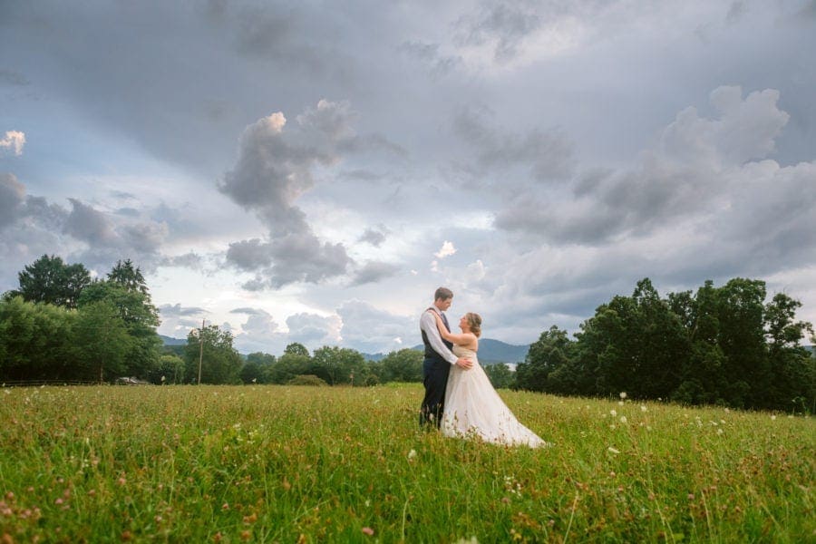 Groom standing in front of bride holding her hips while standing in grassy field smiling at one another
