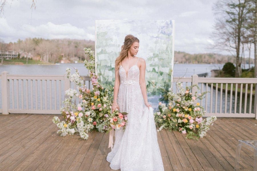 Bride holding bouquet down by side and looking down to the left standing in front of white, green and blue painted canvas backdrop on deck over looking a lake