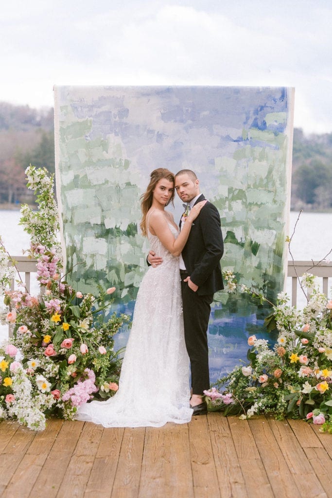 Bride and groom with arms around one another smiling at camera while standing in front of white, green and blue painted canvas backdrop on deck over looking a lake
