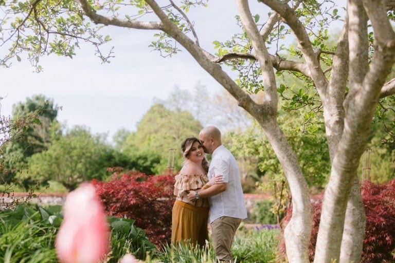 Newly engaged couple hugging one another laughing while standing outside on garden path