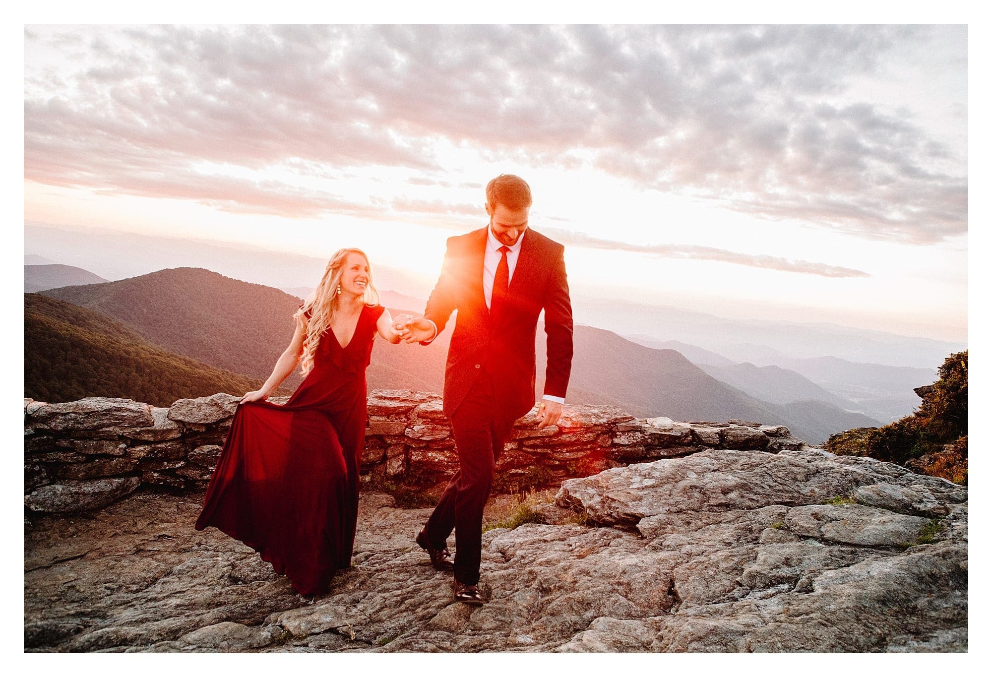 Man in suit holding fiances hand walking along rocky mountain side with mountain range in background at sunset