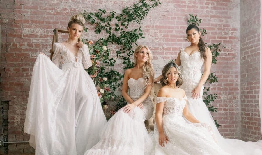 Four women wearing white vintage lace wedding gowns posing in front of light red brick wall with floral greenery running up it