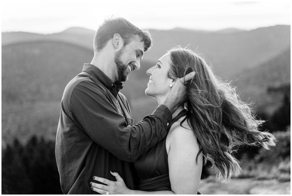 Black and white photo of the couple smiling at each other and the wind blowing her hair on top of the mountain.