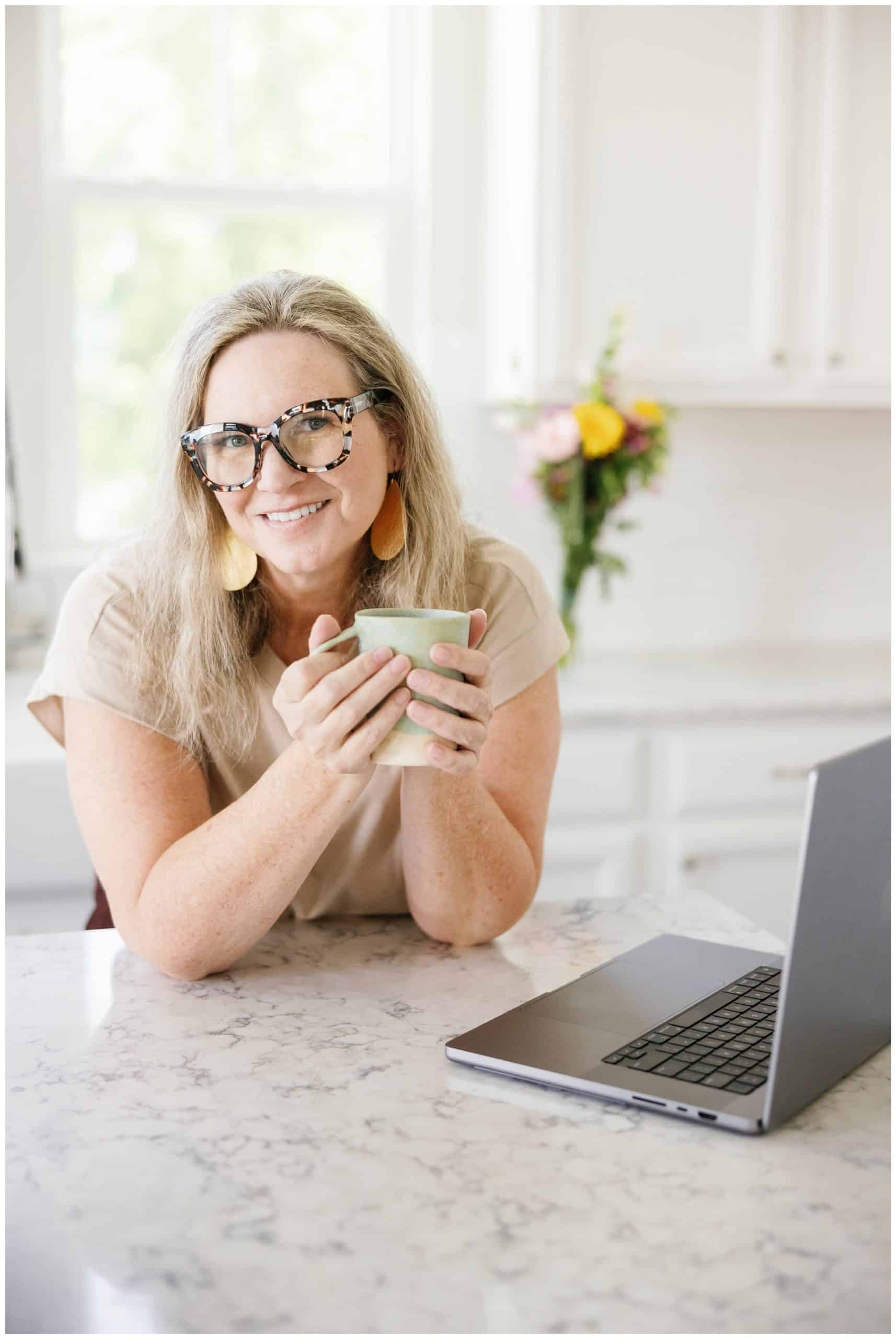 A woman wearing glasses and holding a cup of coffee in front of a laptop.