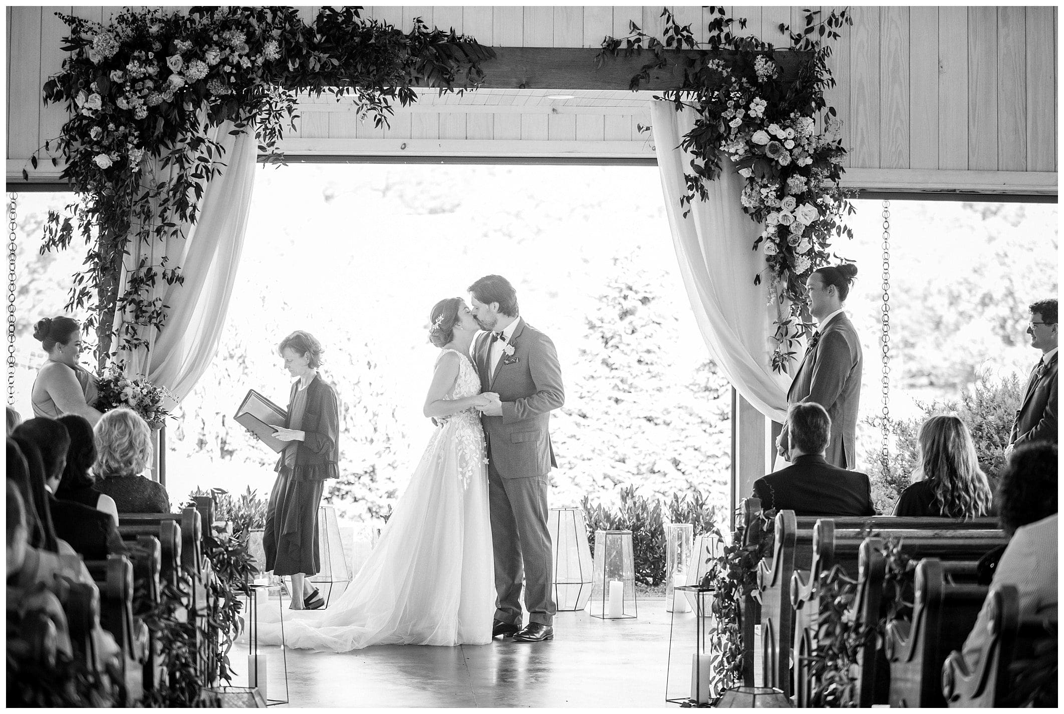 A black and white photo of a bride and groom kissing under an arch.