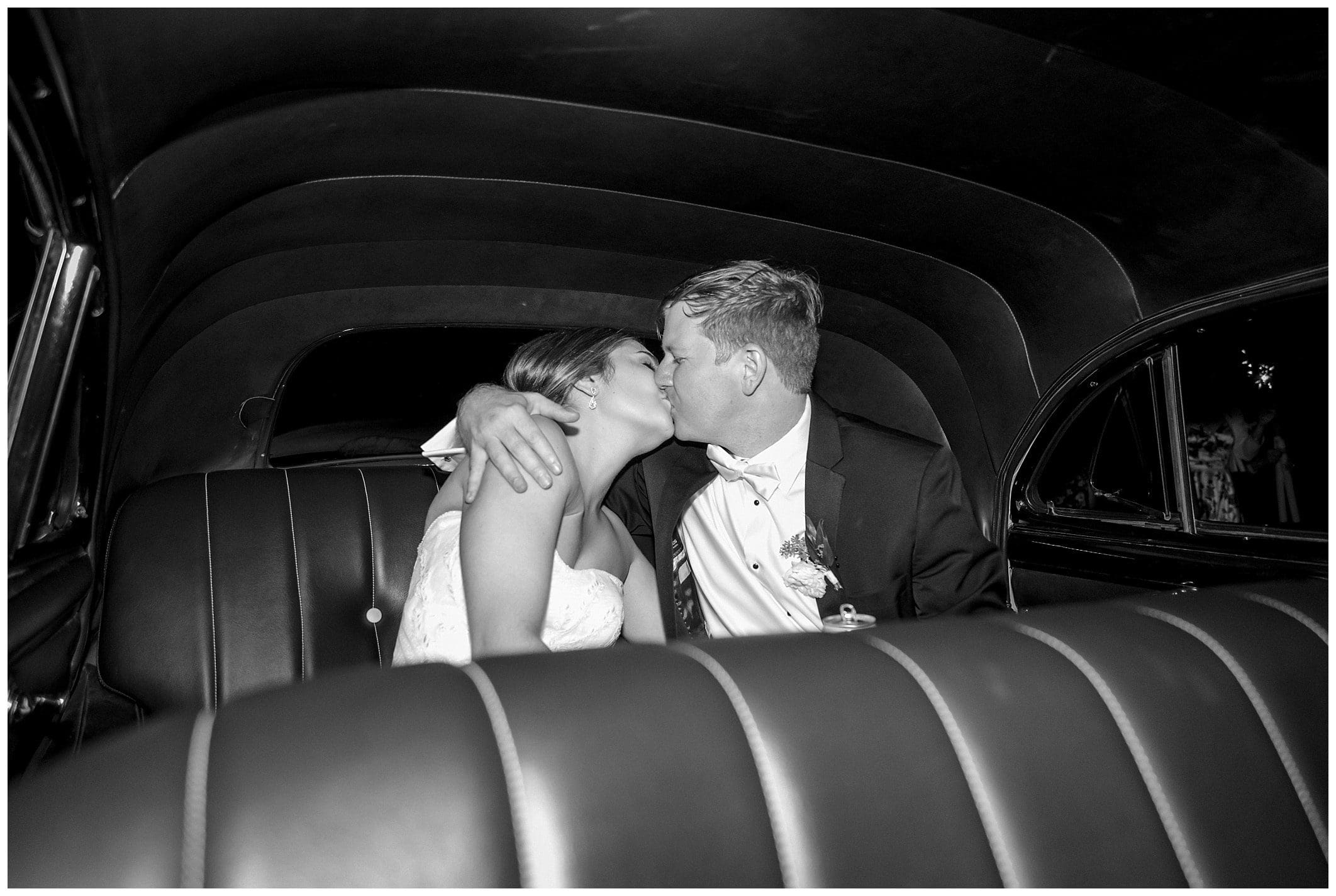 A bride and groom kiss in the back seat of a vintage car.