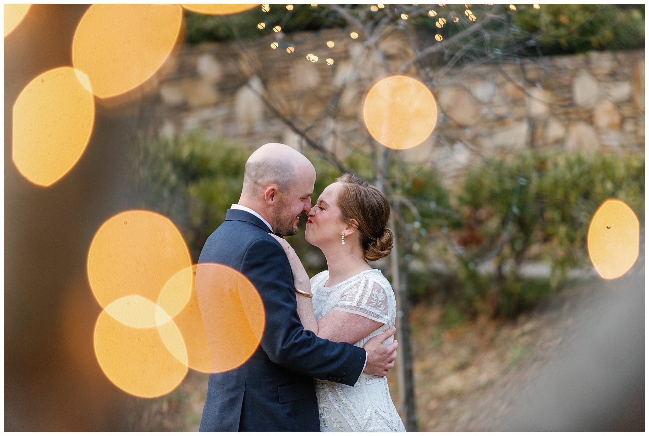 A bride and groom kiss in front of a string of lights.