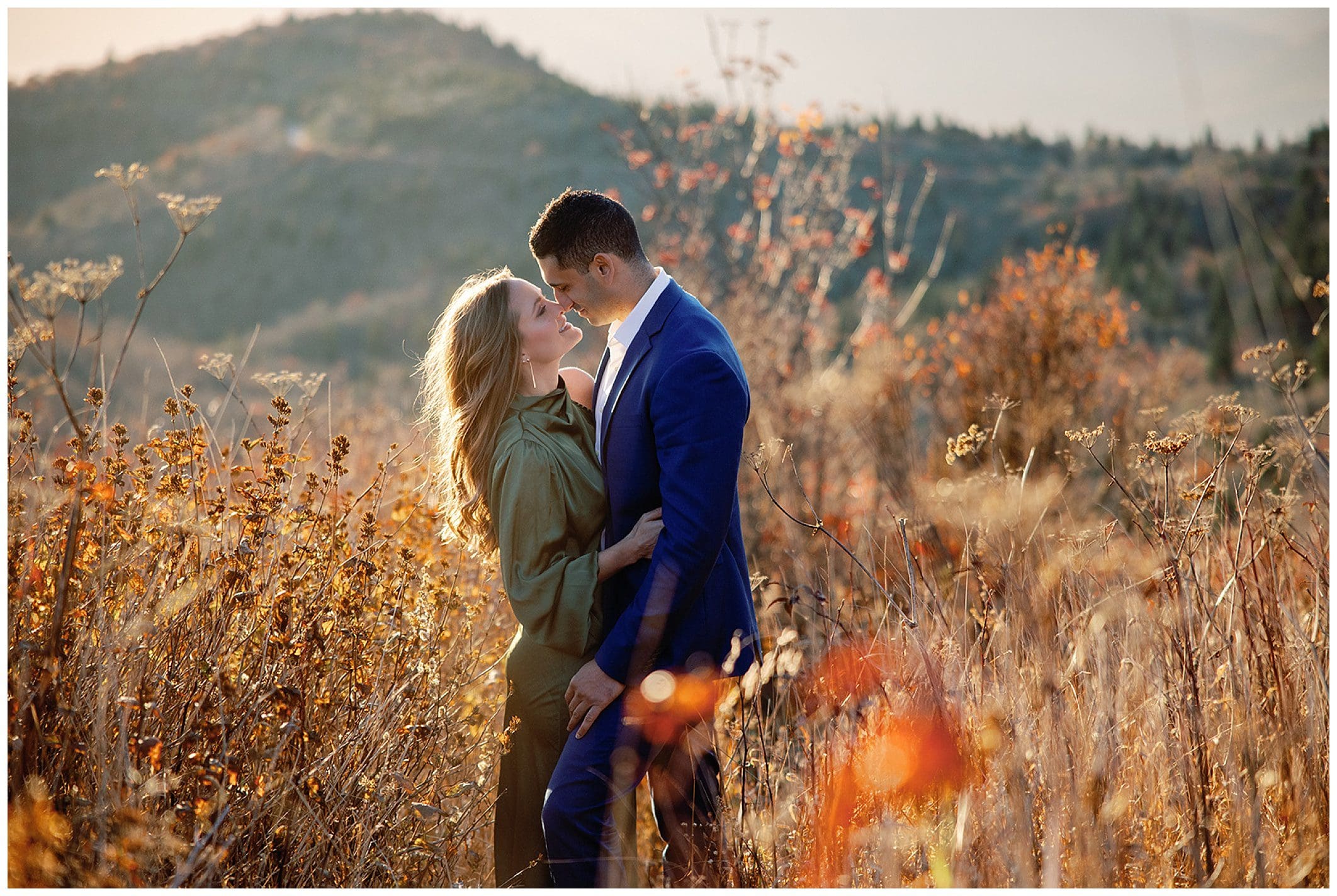 A man in a blue suit and a woman in a green shirt embrace in a sunlit field, celebrating their sunrise engagement.