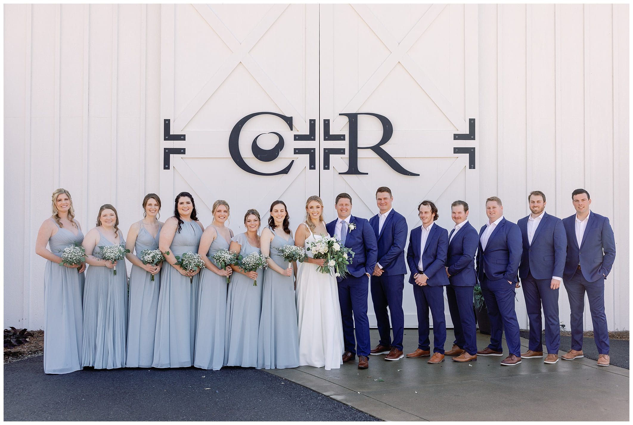 A group of bridesmaids and groomsmen posing in front of a barn.