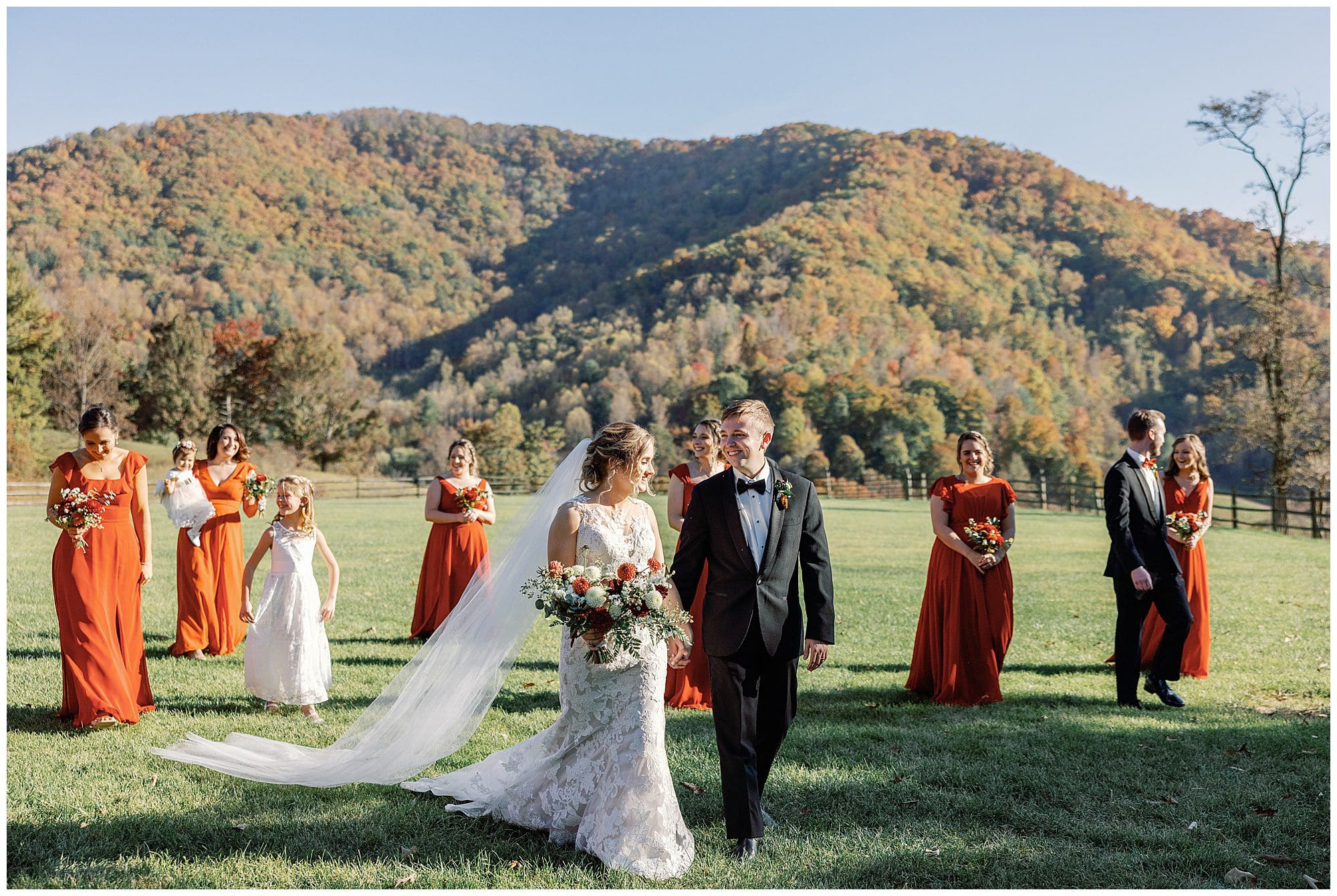 A bride and groom walk hand-in-hand at their outdoor wedding, with bridesmaids in orange dresses and groomsmen in the background, all against a backdrop of autumn mountains.