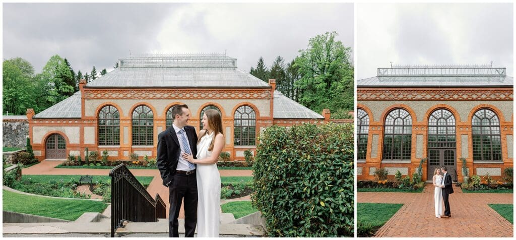 A couple in wedding attire embracing near a brick botanical garden conservatory during their spring engagement session, with a cloudy sky overhead.