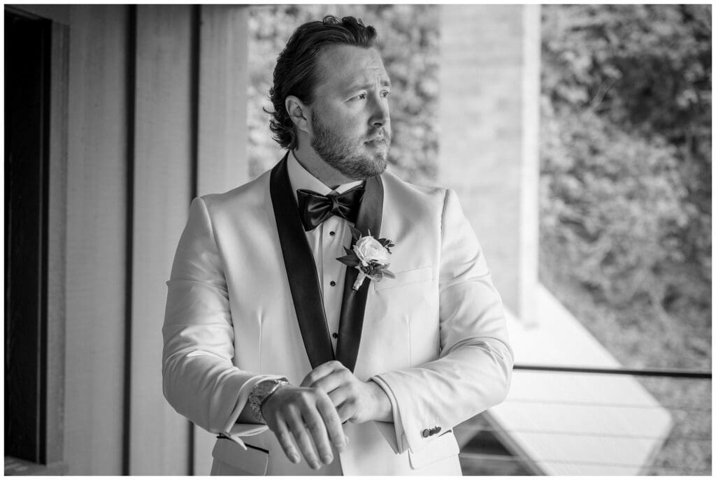 A man in a formal white tuxedo with a bow tie and boutonnière looks pensively out a window in a black and white photograph.