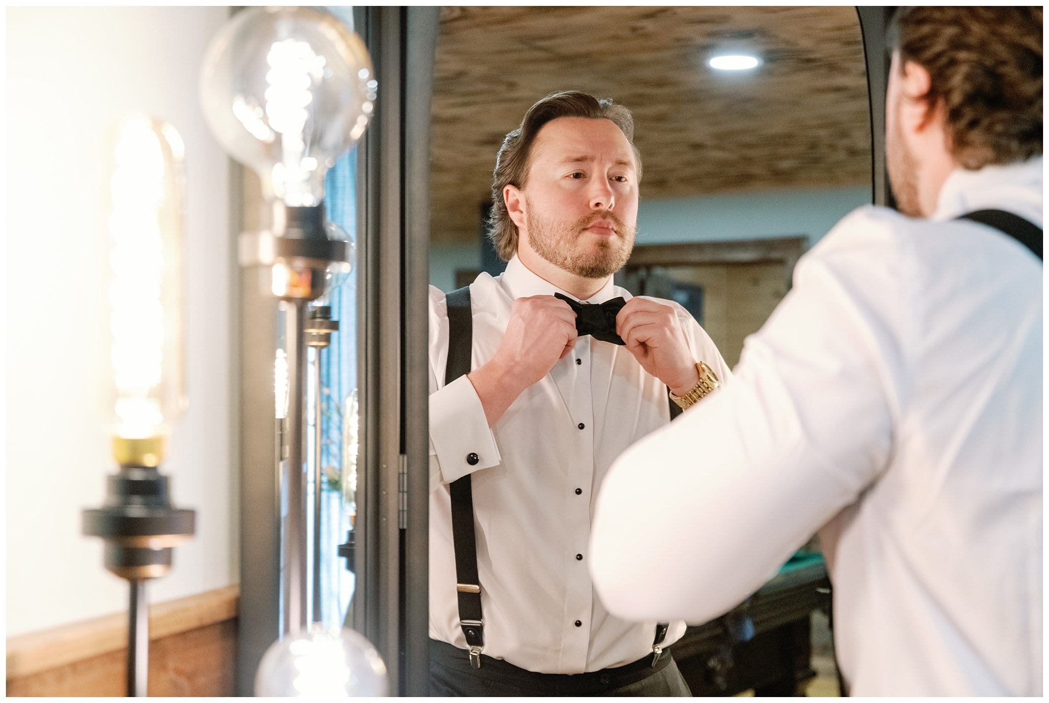 A man in a white dress shirt and suspenders adjusts his bow tie while looking at his reflection in a mirror, surrounded by warm lighting.