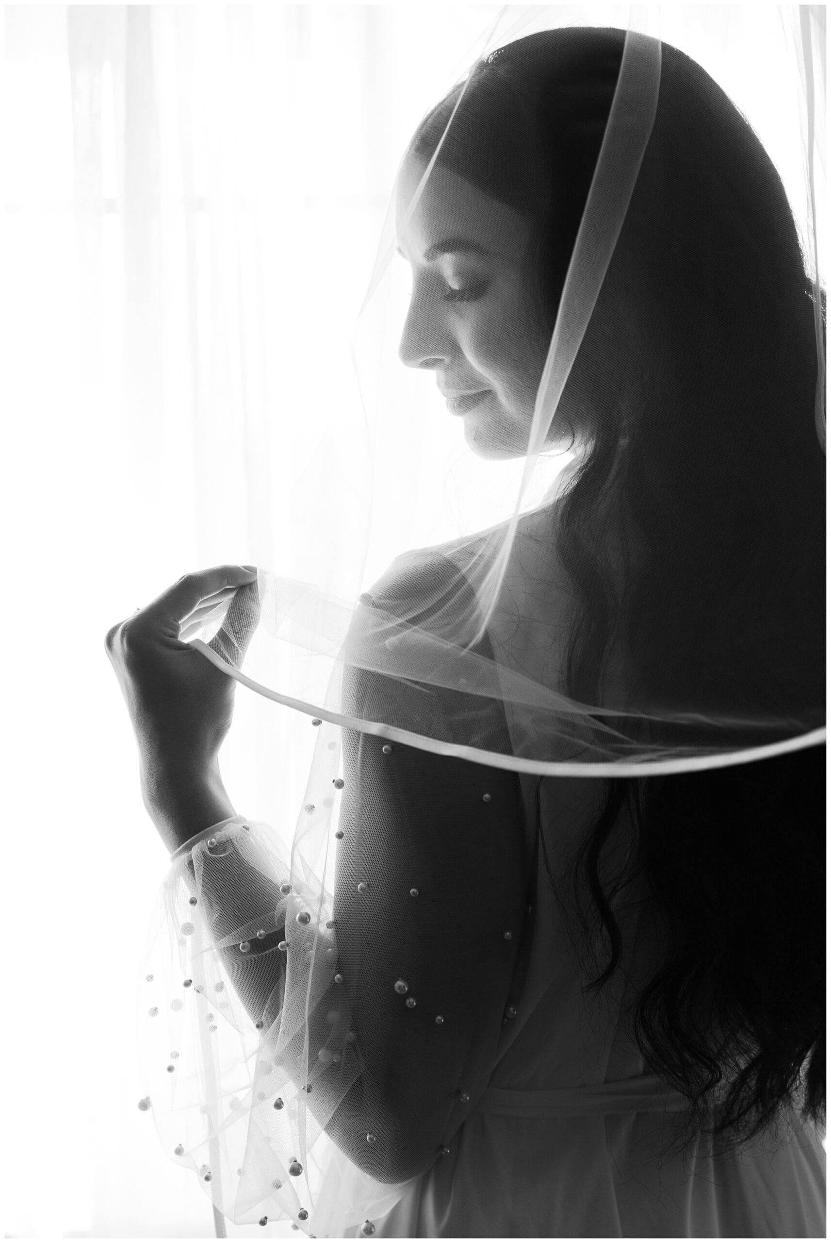 A bride in profile, gently holding her veil, bathed in soft backlight from a large window.