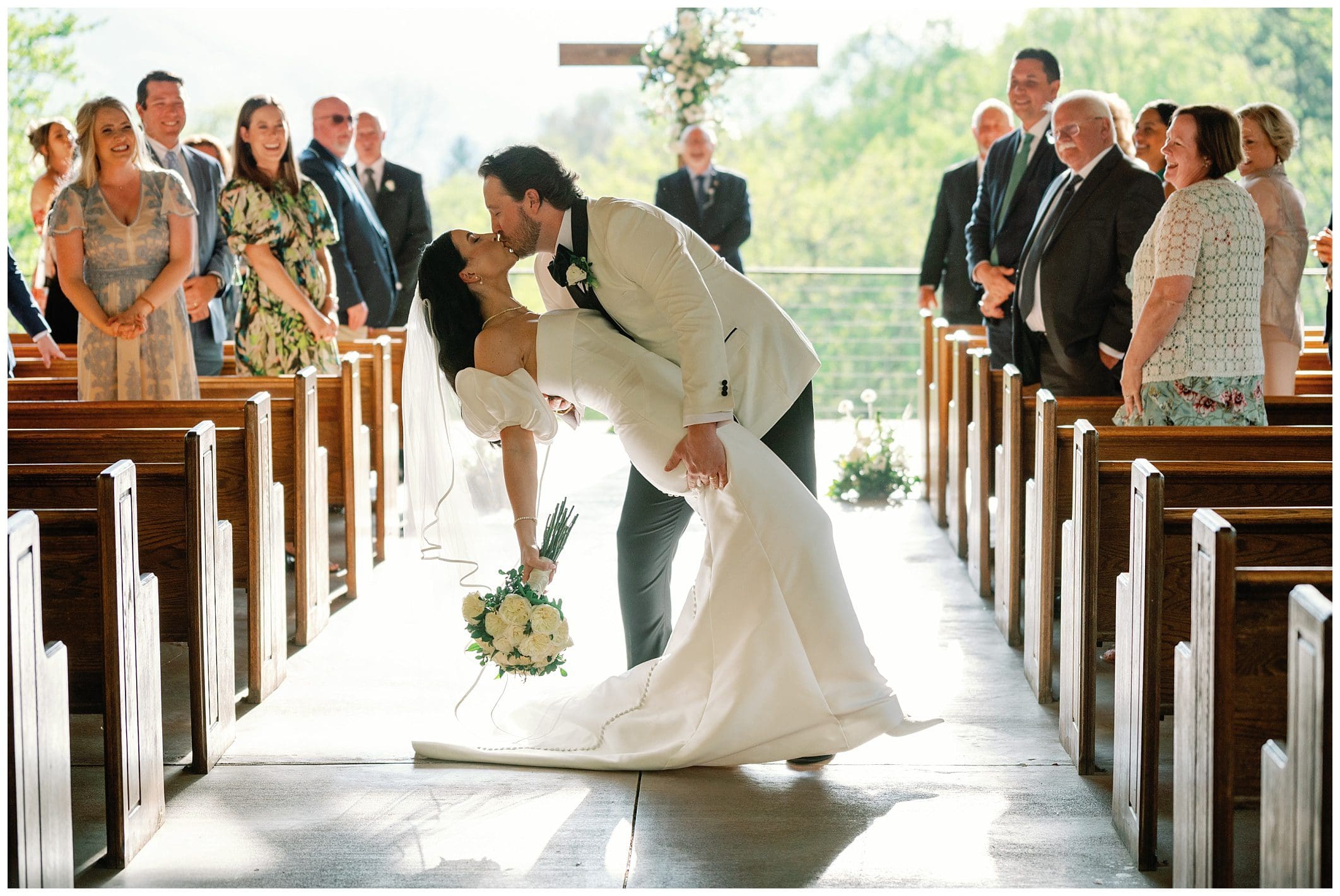 Intimate Spring Microwedding at Parker Mill , bride and groom kiss passionately in a church aisle, surrounded by smiling guests during a sunlit wedding ceremony.