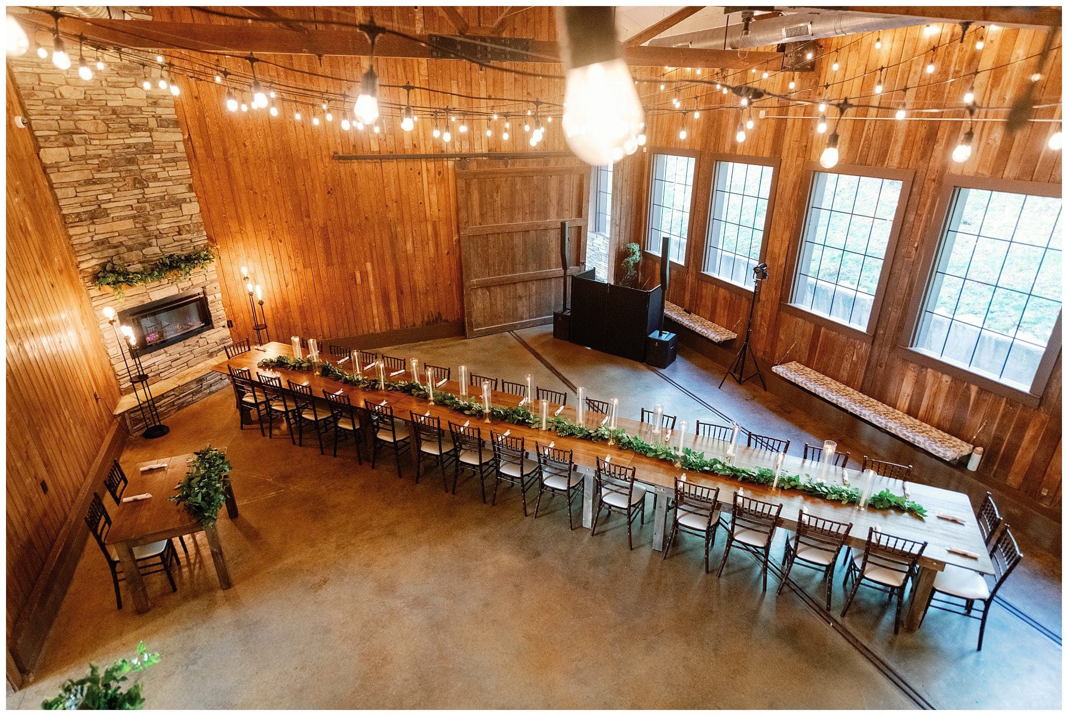 Rustic banquet hall interior with long dining tables set for a meal, string lights overhead, and large windows.