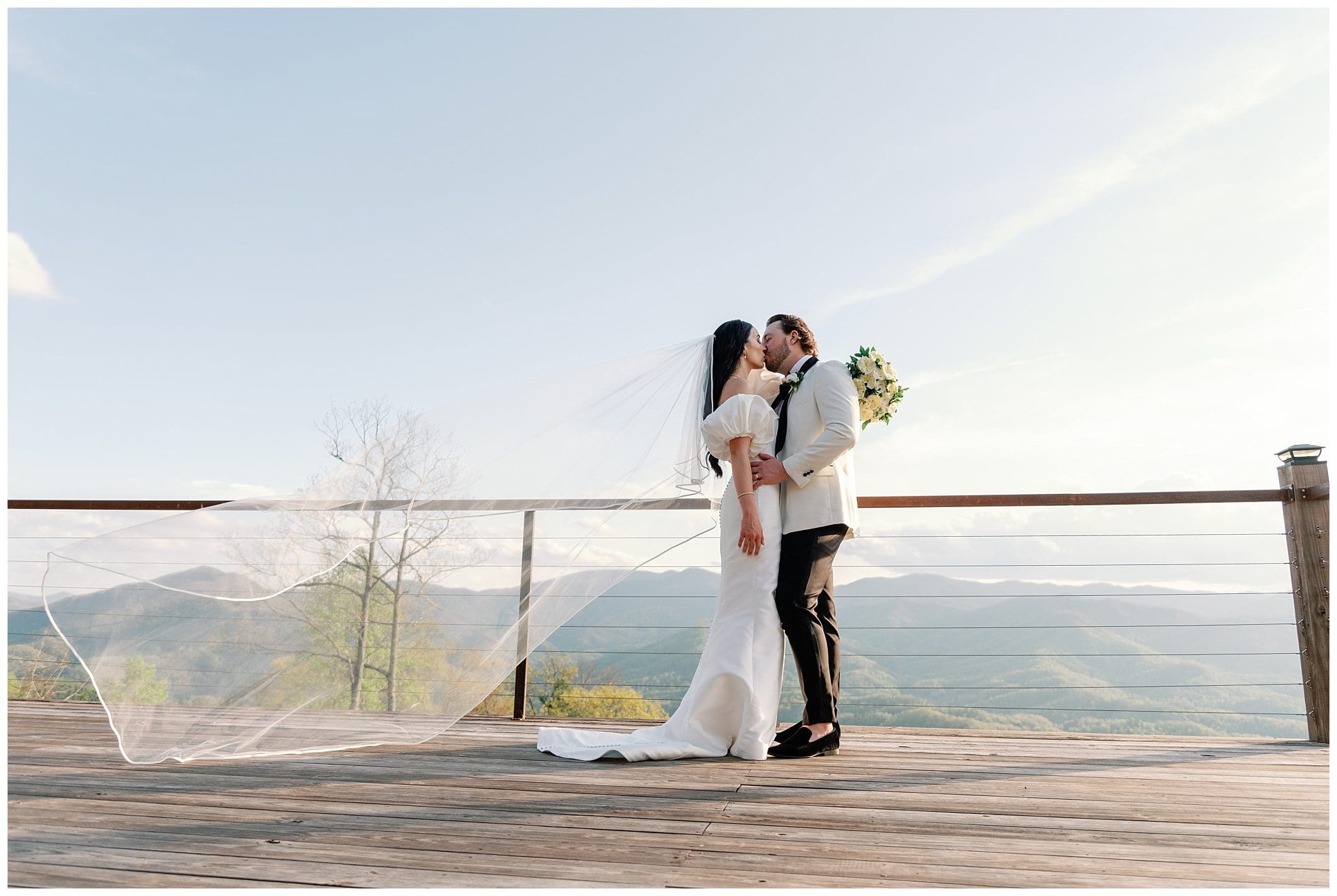 A bride and groom kissing on a deck with a scenic mountain backdrop, the bride's veil flowing in the breeze.