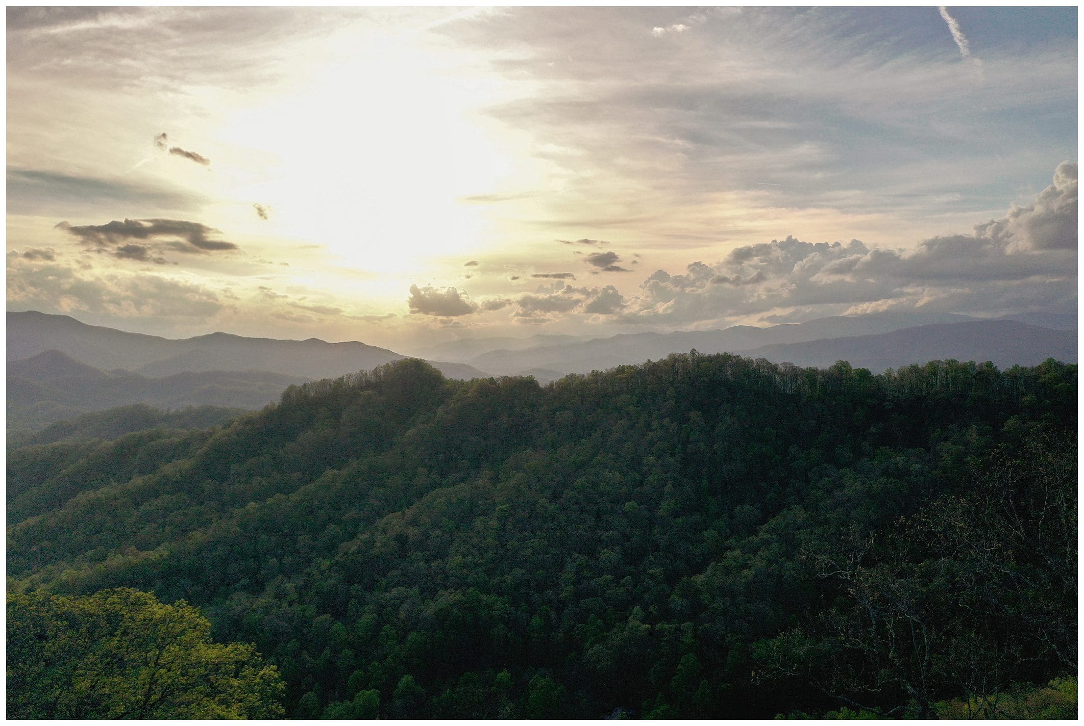 Aerial view of a forest-covered mountain range at sunset, with rays of sunlight breaking through the clouds.