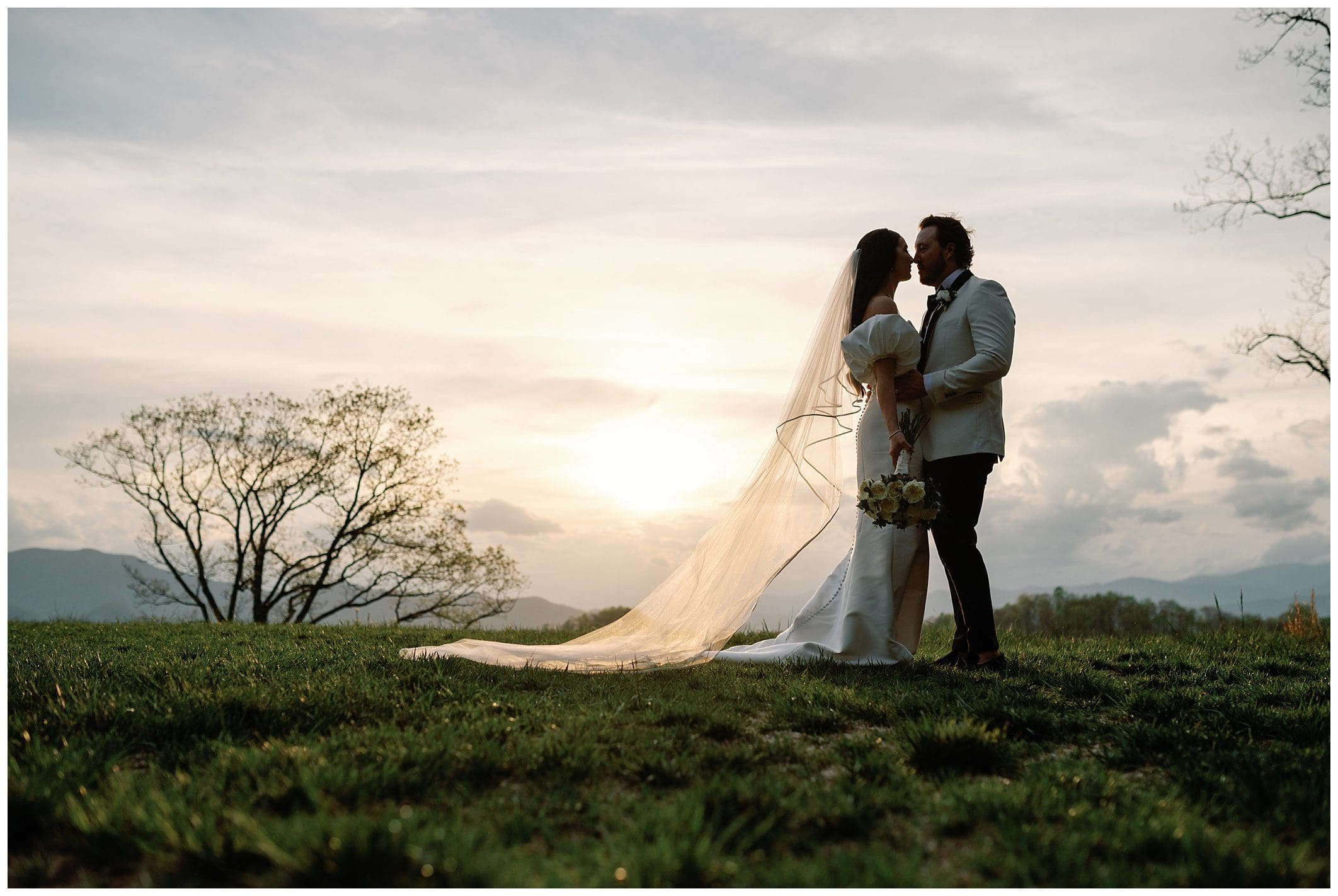 A bride and groom stand close on a grassy hill at sunset, her veil flowing, with mountains and a soft sky in the background.