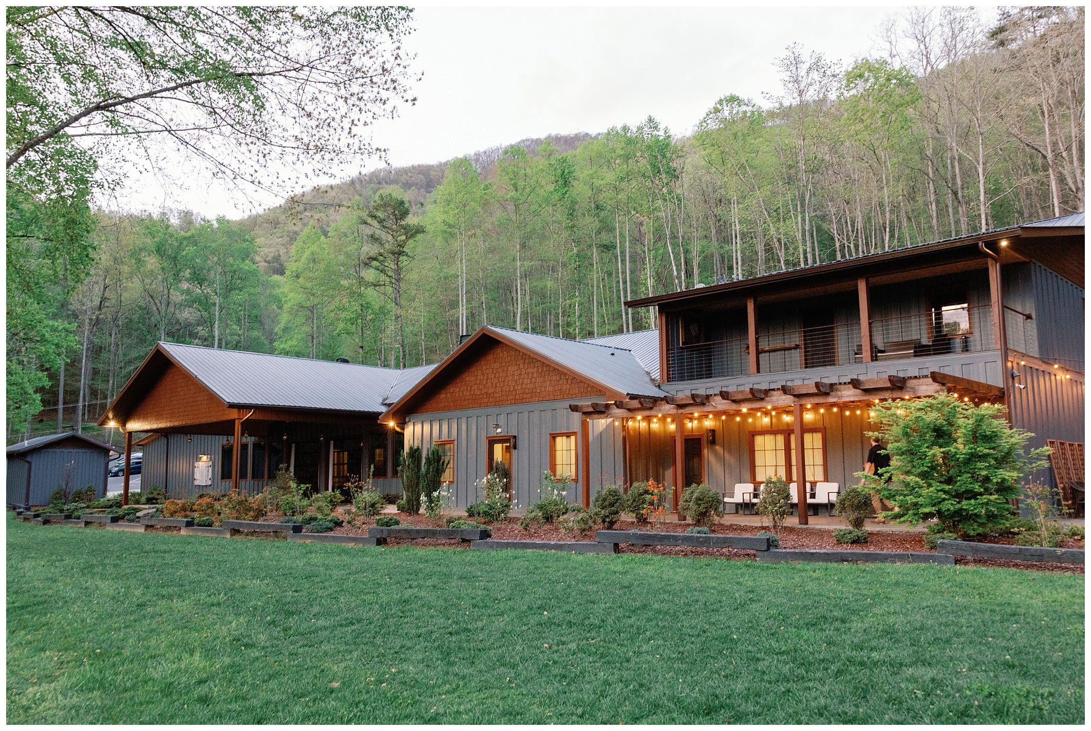 Modern rustic wedding venue with warm lighting, nestled at the base of a lush, tree-covered mountain at dusk.