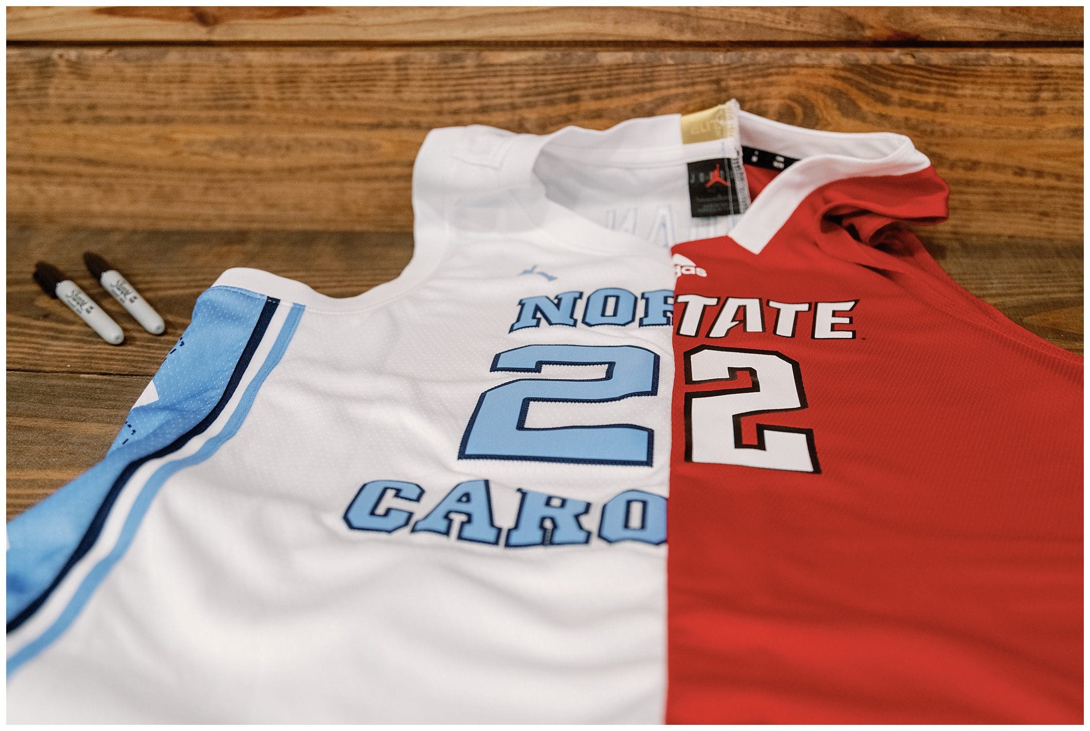 Two basketball jerseys, one white with "north carolina" and the other red with "state," laid on a wooden surface with markers beside them.