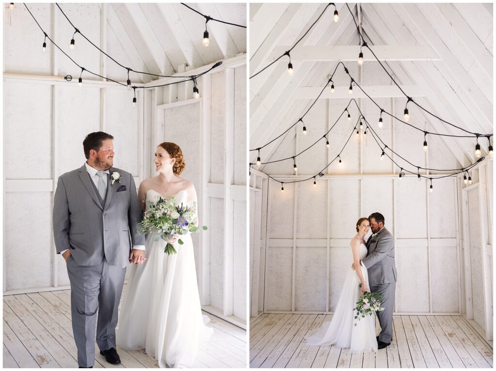 A bride and groom stand in a white wooden room with string lights. The bride wears a white dress and holds a bouquet, while the groom wears a gray suit. They are smiling and embracing each other.