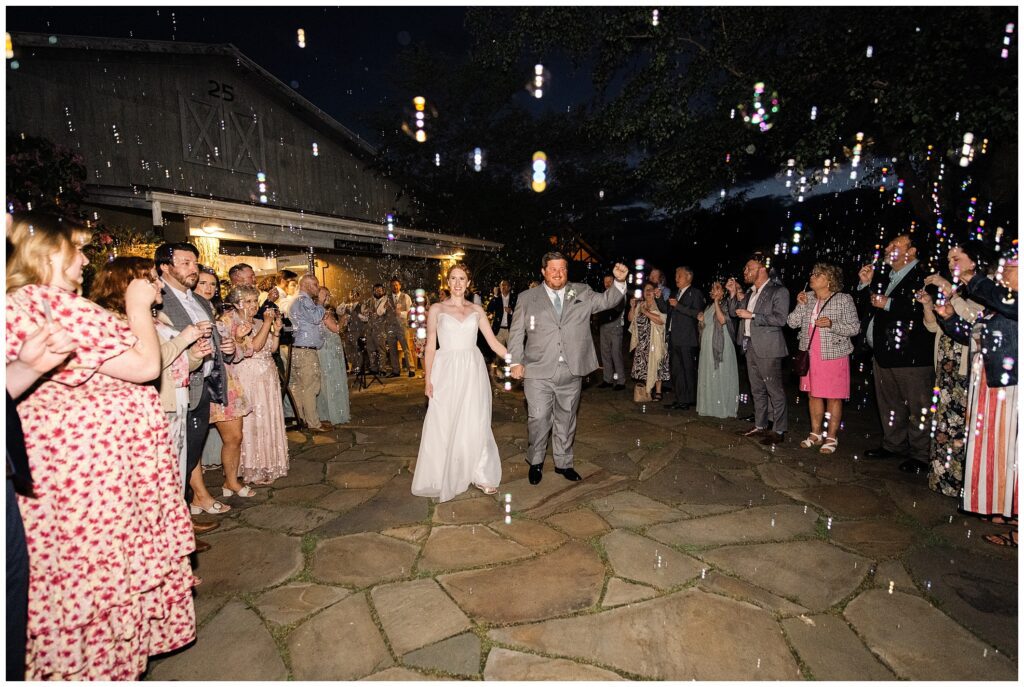 Bride and groom walk hand in hand through a crowd of guests blowing bubbles, outside a barn venue at night.