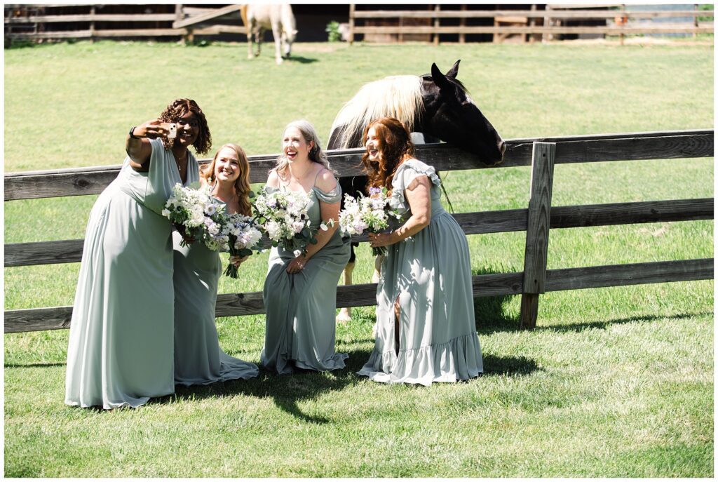 Four bridesmaids in light gray dresses pose and laugh in front of a fence with a black and white horse, holding bouquets of white flowers on a sunny day.