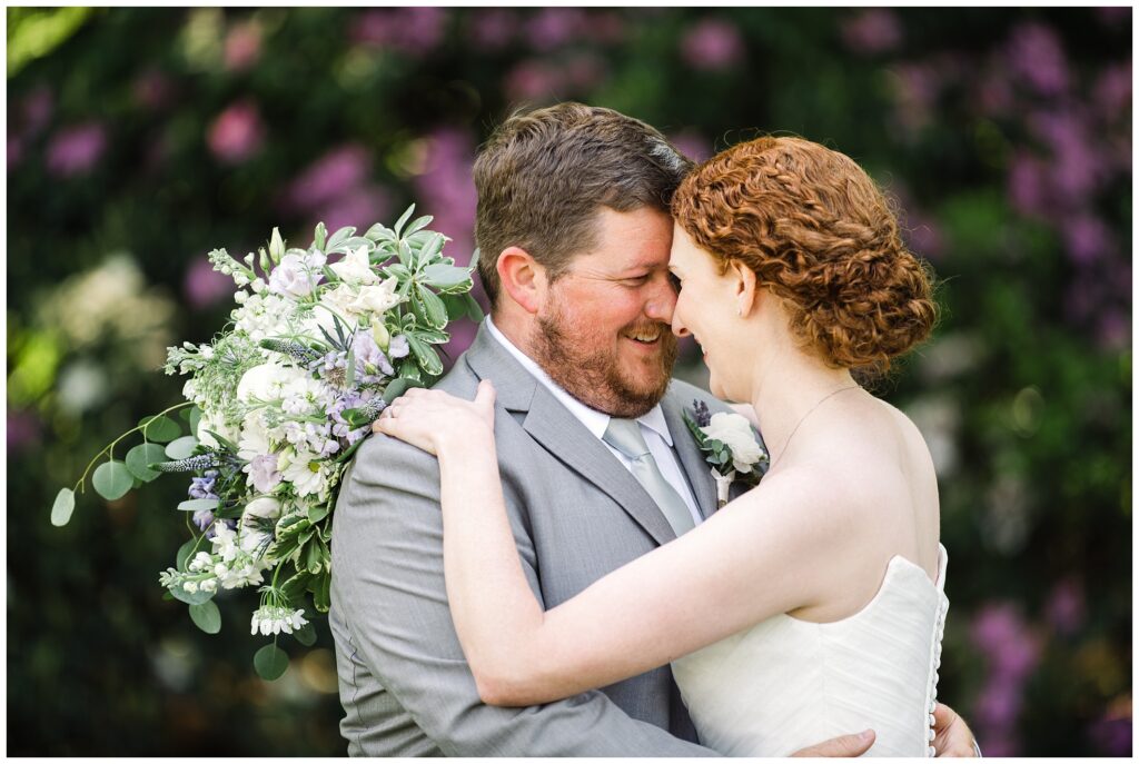 A couple embraces on their wedding day, smiling joyfully. The bride holds a bouquet of white and lavender flowers, and both are dressed in formal attire. Greenery and flowers are in the background.