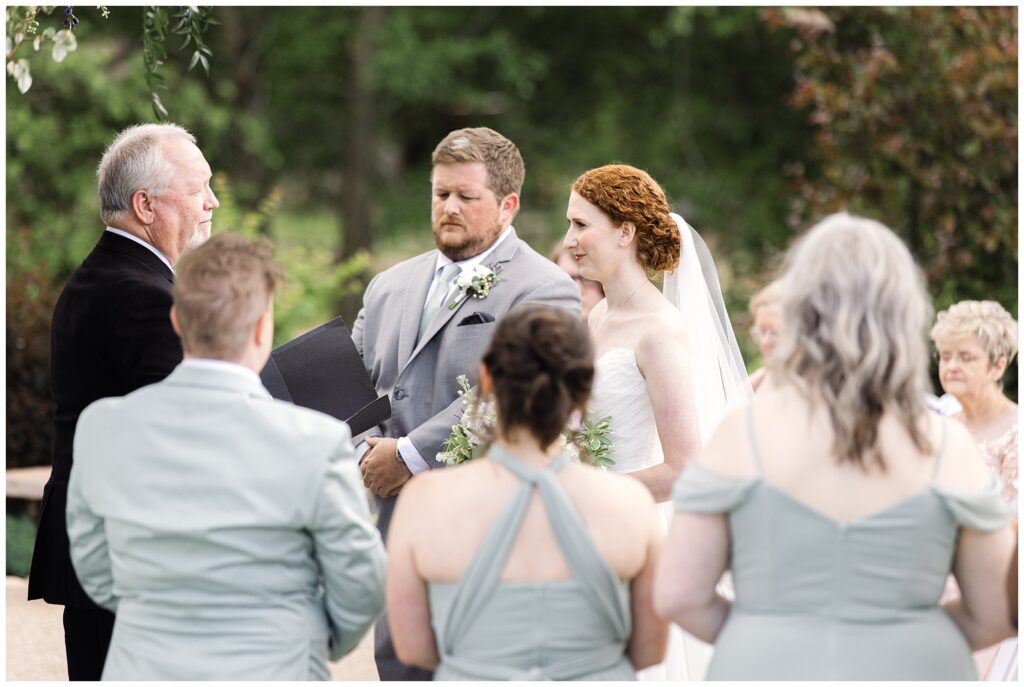 A couple exchanging vows outdoors in a wedding ceremony, surrounded by their bridal party in light grey attire, with a forested background.