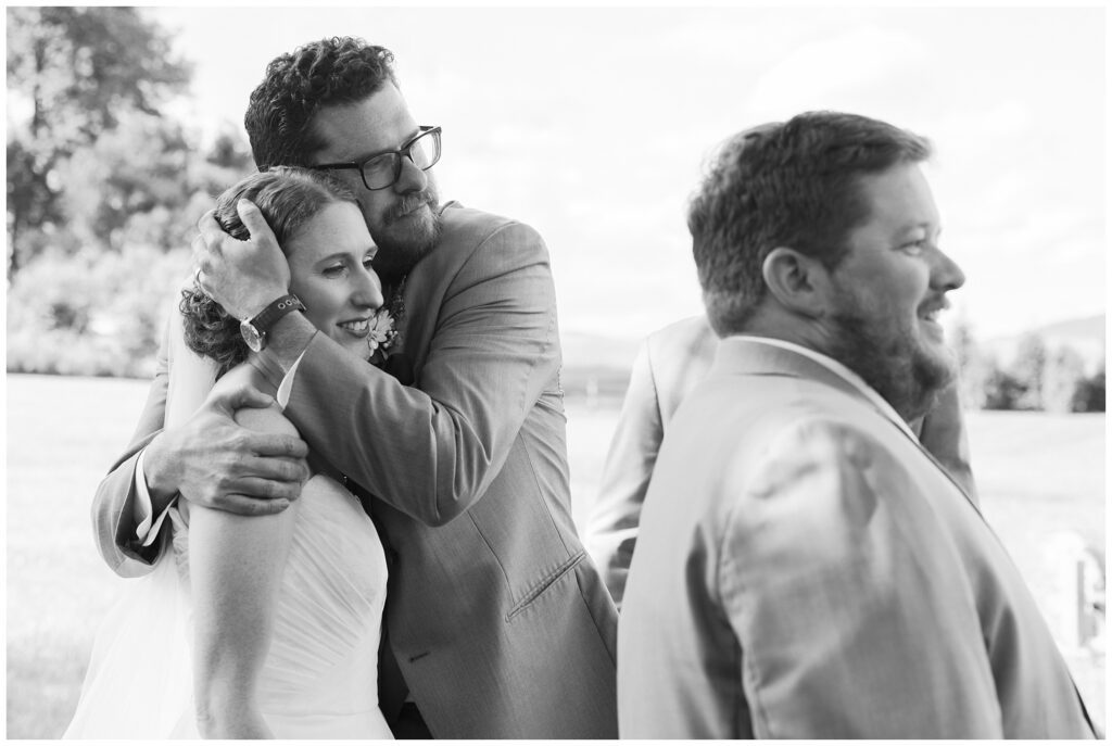 A bride and groom share an embrace while a man in a suit stands nearby outdoors. The scene appears to be at a wedding.