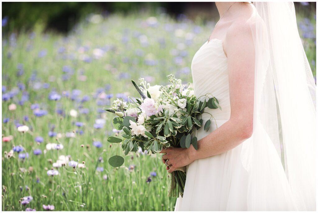 A bride in a white dress holding a bouquet of flowers, standing in a field of wildflowers. The bride's face is not visible in the image.