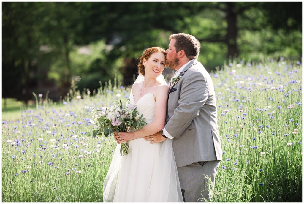 A bride and groom embrace in a field of wildflowers, with the groom smiling and the bride holding a bouquet, both wearing wedding attire.