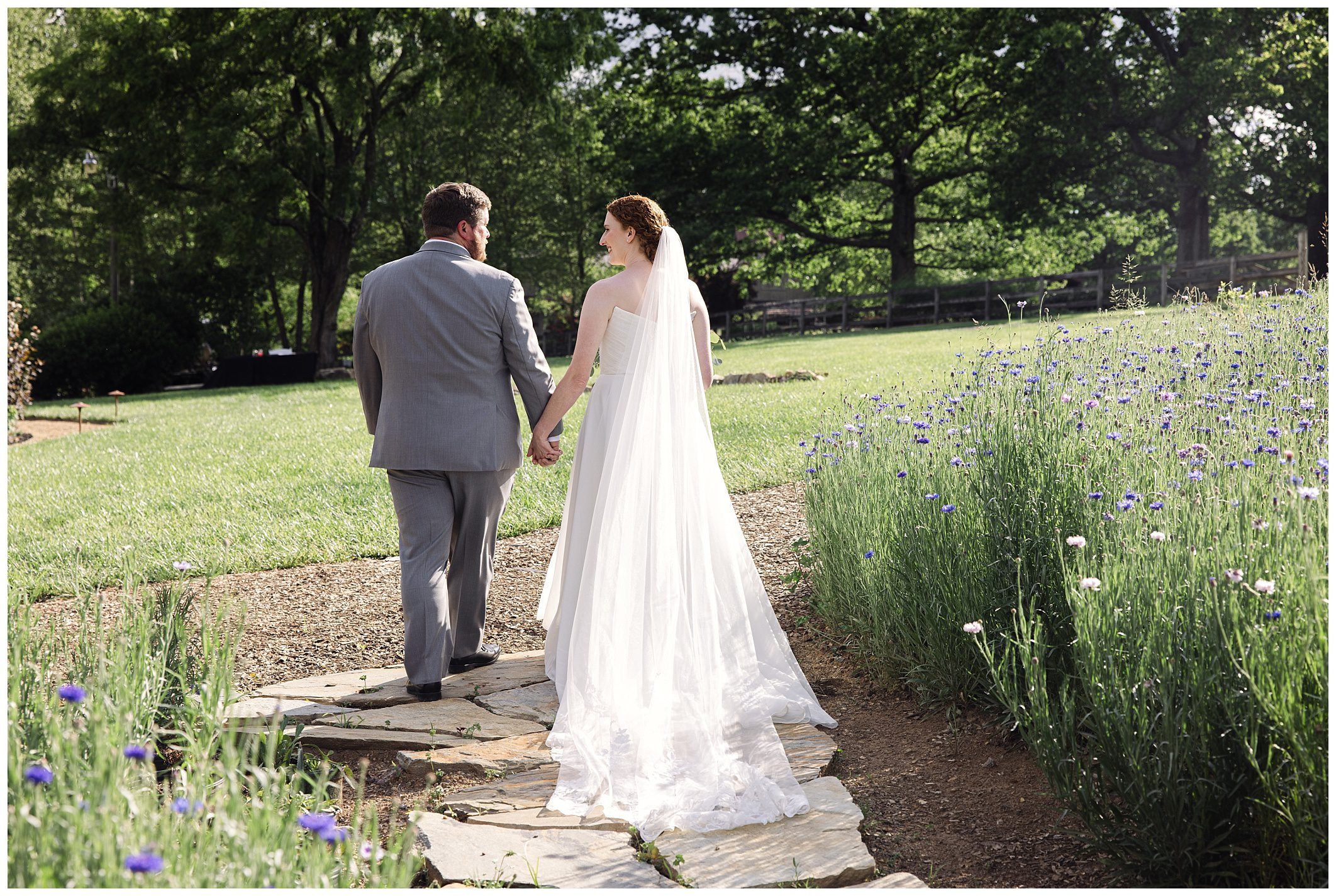 A bride in a white dress and veil, and a groom in a gray suit, walk hand in hand along a stone path in a garden surrounded by greenery and flowers.