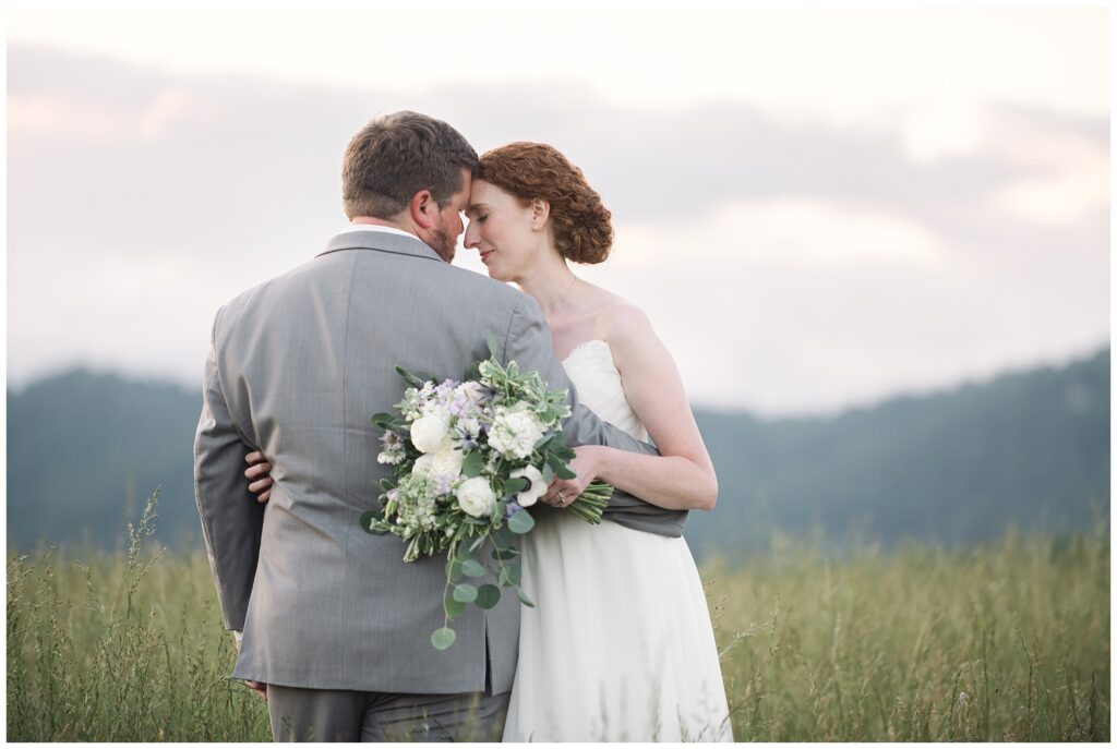 A bride and groom are standing closely, holding a bouquet of flowers, in an outdoor setting with grass and hills in the background.