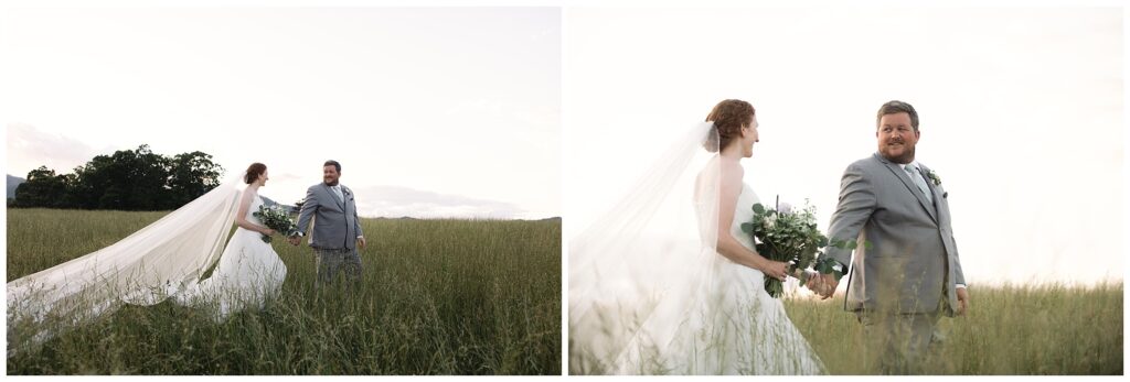 A couple in wedding attire walk through a field. The bride wears a white dress with a long veil and holds a bouquet, while the groom wears a gray suit.