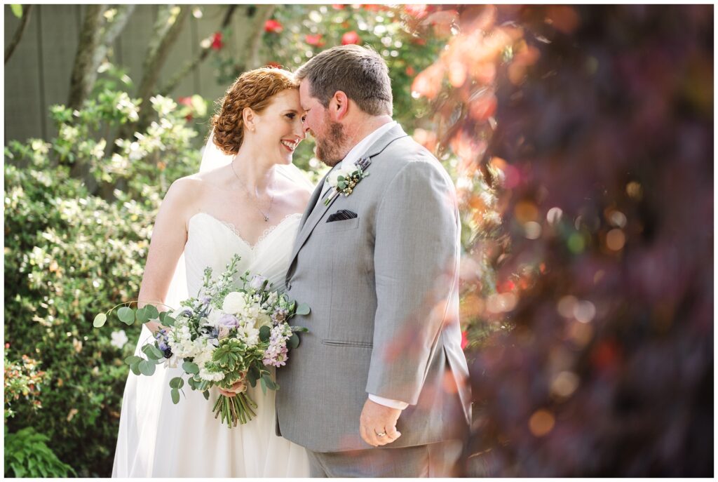 A bride in a white dress and a groom in a gray suit share a moment, standing closely together in an outdoor, greenery-filled setting. The bride holds a bouquet of flowers.