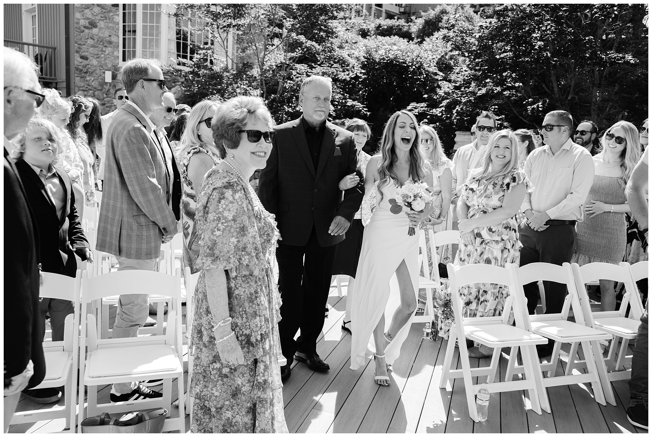 A bride walks down the aisle with an older man, surrounded by seated and standing guests, at an outdoor wedding.