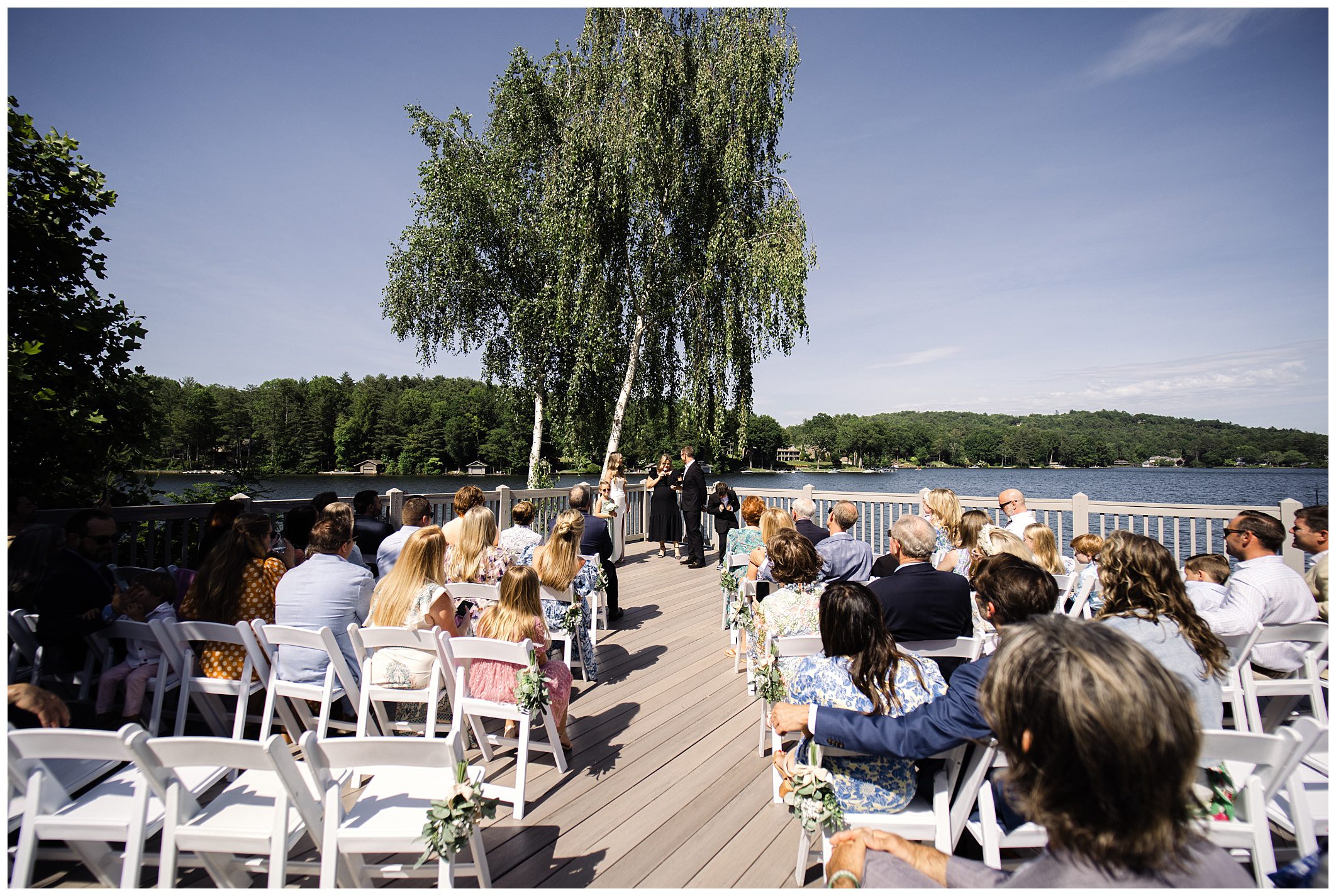 A wedding ceremony taking place outdoors on a wooden deck by a lake, with guests seated in rows of white chairs and the couple standing under a large tree.