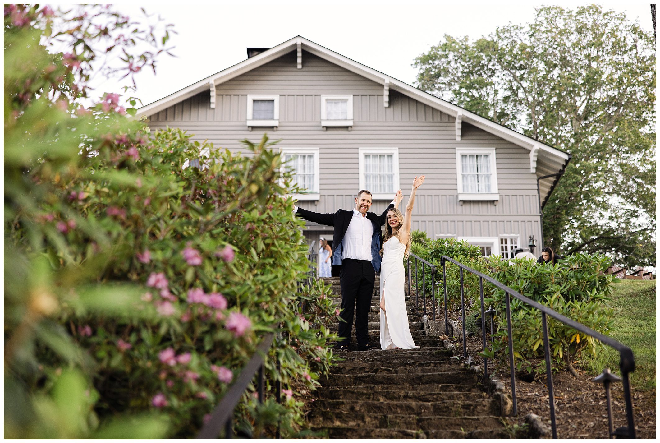 A couple stands on outdoor steps, smiling and waving. The man wears a suit, the woman a white dress. They are in front of a grey house with large windows and surrounded by greenery and flowers.