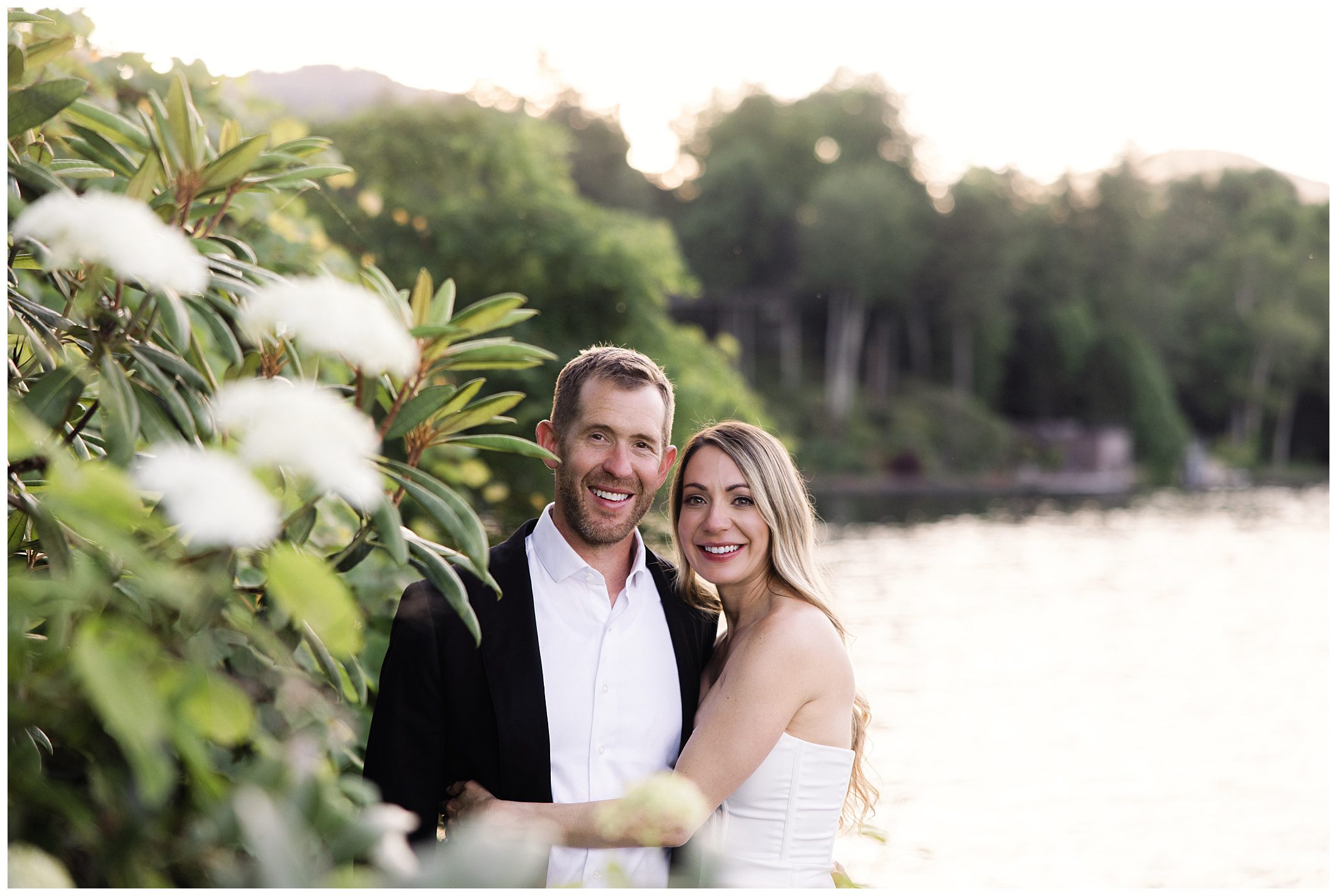 A man and woman stand close together smiling, with a scenic background of a lake and greenery.