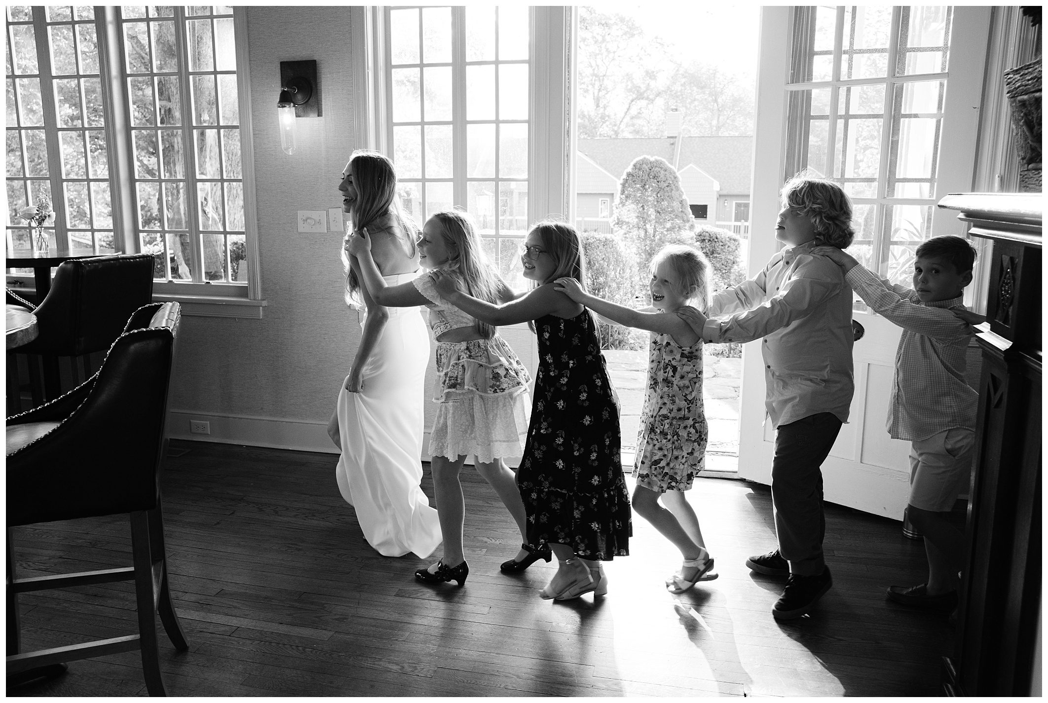 Black and white photo of a group of children and a woman in a white dress, lined up and holding each other's shoulders, walking in a well-lit room with large windows.