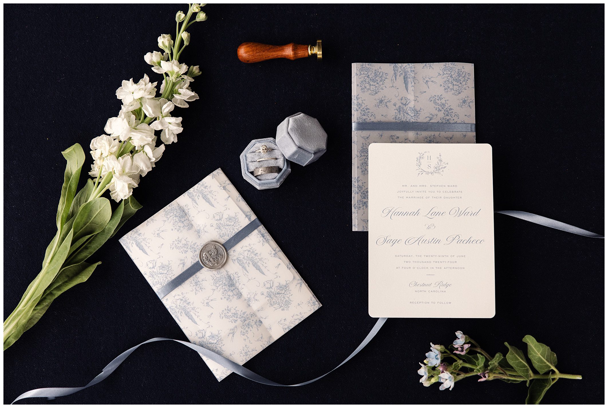 Elegant wedding invitation suite with floral designs, a wax seal, a ribbon, a white flower, and a box containing two rings. All items are arranged on a dark background.