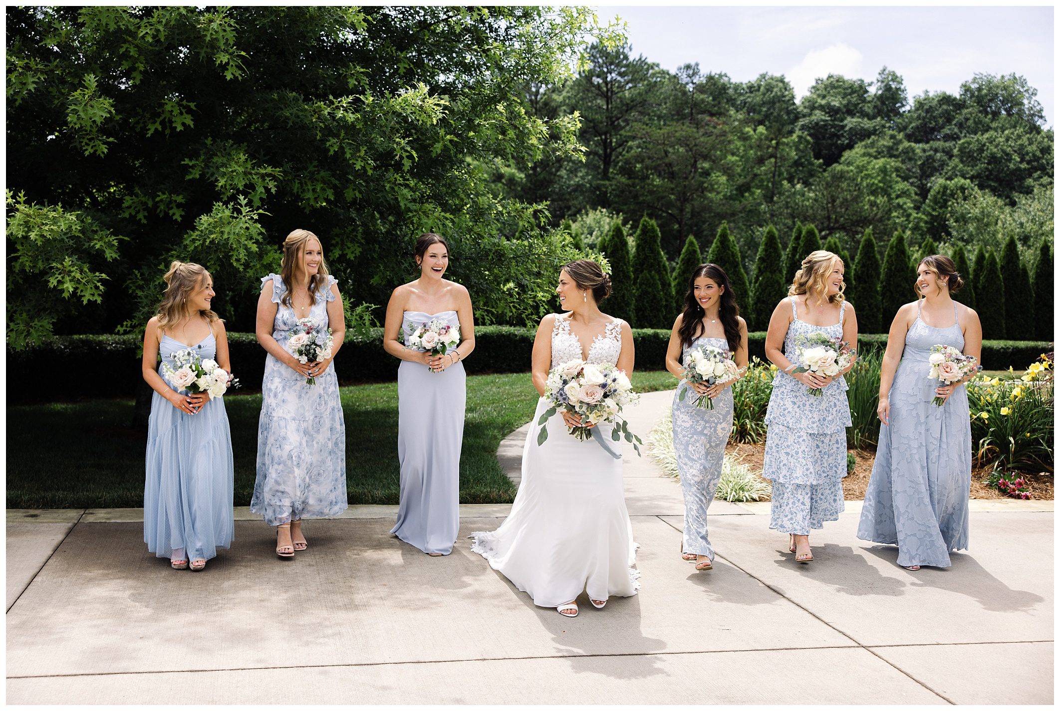 A bride in a white dress stands with six bridesmaids in light blue dresses, all holding bouquets at a summer wedding in Asheville. They are outdoors on a paved walkway with greenery in the background.